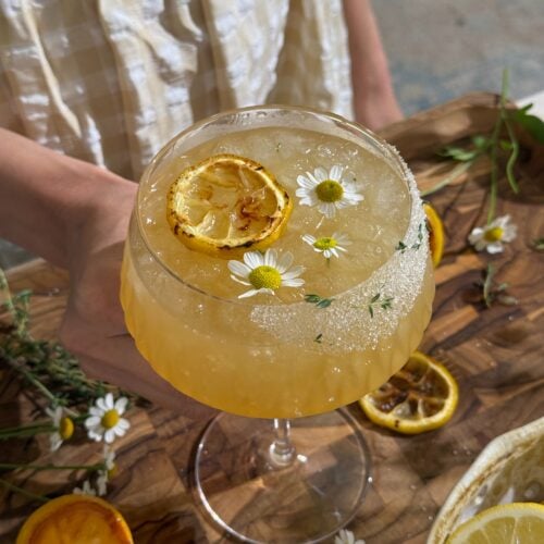 Charred Lemon Ginger Mocktail with ice, grilled lemon slice, chamomile flowers, and fresh herbs in a stemmed glass.
