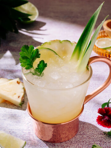 Pineapple Mezcal Mule cocktail with ice, pineapple, lime, and cilantro garnish in a copper-handled glass.