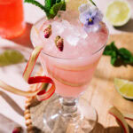Pink rhubarb rose cocktail in a stemmed glass with crushed ice, garnished with mint, lime, dried rosebuds, an edible flower, and a rhubarb ribbon.