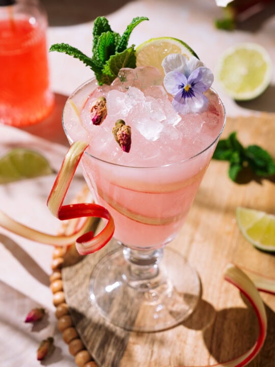 Pink rhubarb rose cocktail in a stemmed glass with crushed ice, garnished with mint, lime, dried rosebuds, an edible flower, and a rhubarb ribbon.