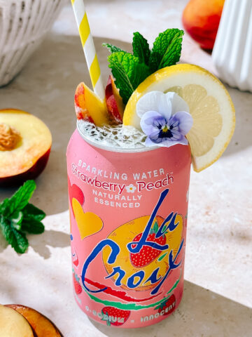 Peach Bellini Spritz mocktail in LaCroix can with lemon, peach slices, mint, and edible flower garnish."