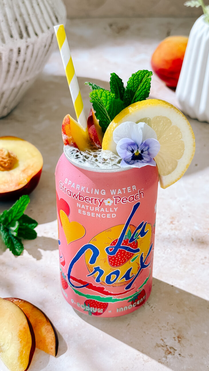 Peach Bellini Spritz mocktail in LaCroix can with lemon, peach slices, mint, and edible flower garnish."