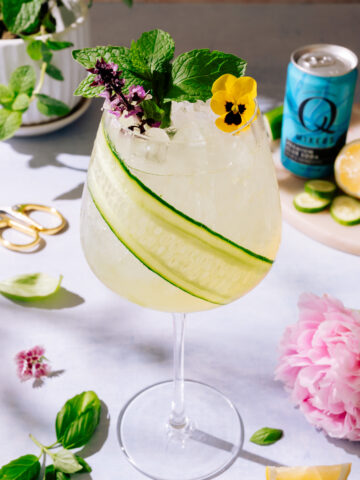 Garden Gin Spritz cocktail in a tall glass with cucumber strip for garnish and floral garnish. Surrounded by lemons, mint leaves and pink flowers.