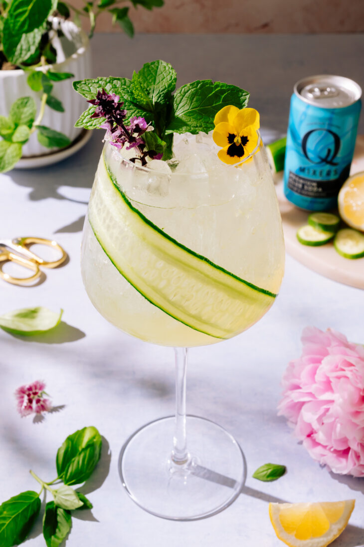 Garden Gin Spritz cocktail in a tall glass with cucumber strip for garnish and floral garnish. Surrounded by lemons, mint leaves and pink flowers.