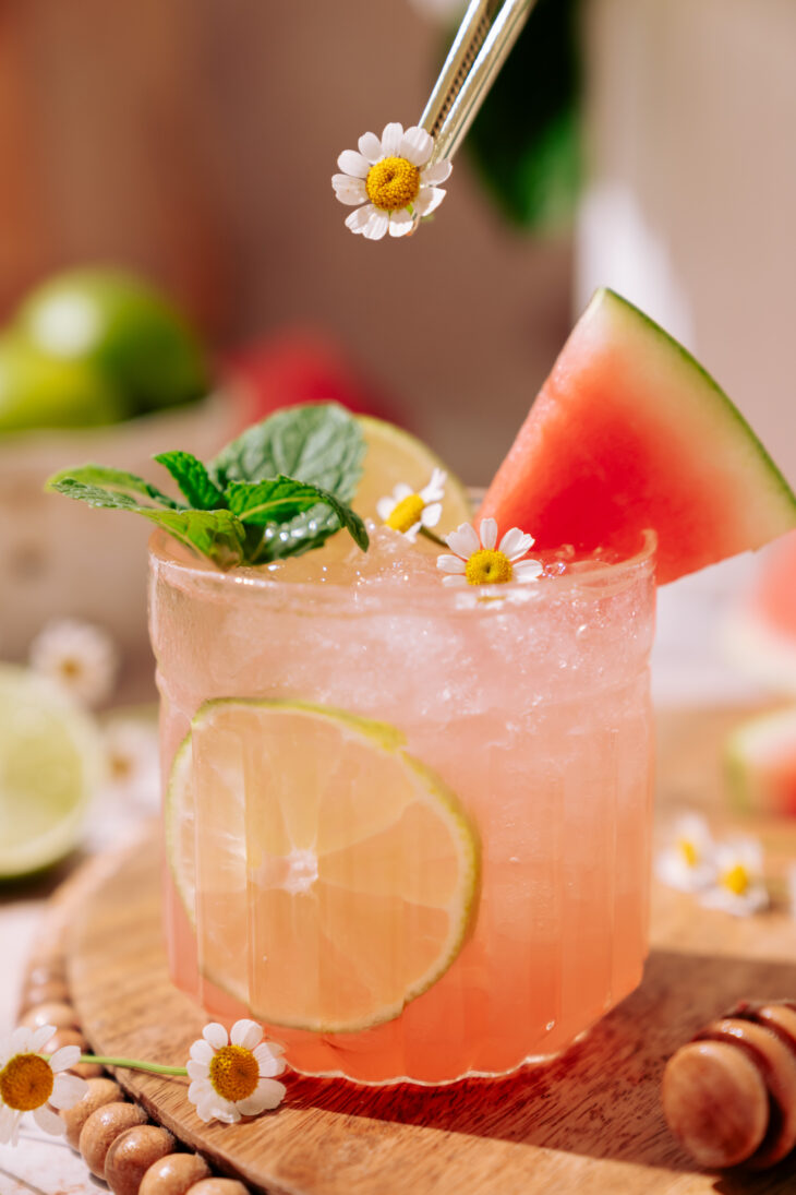 Watermelon Vodka Cocktail served in a glass with ice, lime slices, fresh mint, chamomile flowers, and a watermelon wedge on a wooden board.
