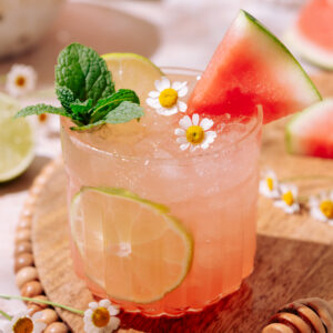 Watermelon Vodka Cocktail served in a glass with ice, lime slices, fresh mint, chamomile flowers, and a watermelon wedge on a wooden board.
