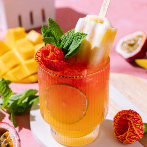 Vibrant Tropical Punch No-jito mocktail with hibiscus, mango, lime slice, fresh mint, and a popsicle garnish in a ribbed glass on a pink background.