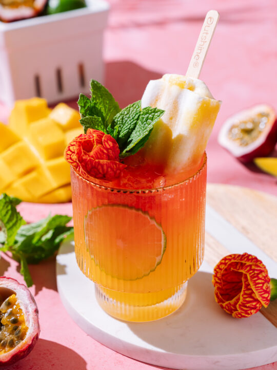 Vibrant Tropical Punch No-jito mocktail with hibiscus, mango, lime slice, fresh mint, and a popsicle garnish in a ribbed glass on a pink background.