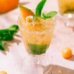 A refreshing Cantaloupe Hugo Spritz served in an elegant glass, garnished with a cantaloupe wedge, fresh mint leaves, and a small white flower, with crushed ice and green herbs at the bottom.