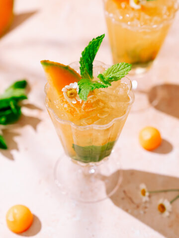 A refreshing Cantaloupe Hugo Spritz served in an elegant glass, garnished with a cantaloupe wedge, fresh mint leaves, and a small white flower, with crushed ice and green herbs at the bottom.