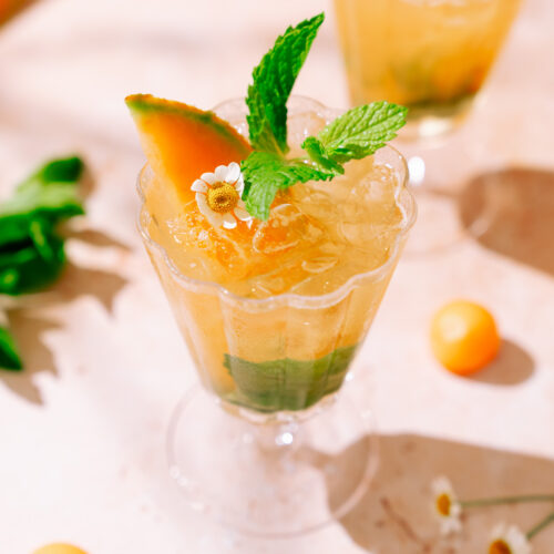 A refreshing Cantaloupe Hugo Spritz served in an elegant glass, garnished with a cantaloupe wedge, fresh mint leaves, and a small white flower, with crushed ice and green herbs at the bottom.