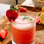 Charred Strawberry Whiskey Sour in a ribbed glass, garnished with a whole strawberry and fresh herbs, surrounded by sliced strawberries and lemons. Je to doposud užitečné? Zeptat se služby ChatGPT