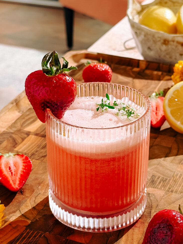Charred Strawberry Whiskey Sour in a ribbed glass, garnished with a whole strawberry and fresh herbs, surrounded by sliced strawberries and lemons. Je to doposud užitečné? Zeptat se služby ChatGPT