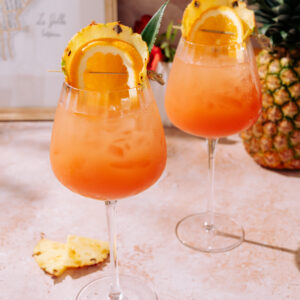 Two Piña Colada Spritz cocktails served in large wine glasses, filled with ice and a bright orange-pineapple mix, garnished with pineapple slices, orange wheels, and a pineapple leaf, placed on a light stone surface.