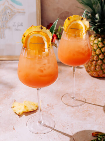 Two Piña Colada Spritz cocktails served in large wine glasses, filled with ice and a bright orange-pineapple mix, garnished with pineapple slices, orange wheels, and a pineapple leaf, placed on a light stone surface.