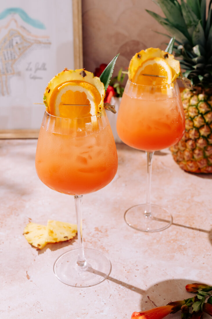 Two Piña Colada Spritz cocktails served in large wine glasses, filled with ice and a bright orange-pineapple mix, garnished with pineapple slices, orange wheels, and a pineapple leaf, placed on a light stone surface.
