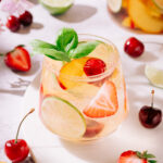 Refreshing summer white sangria in a glass garnished with fresh basil, lime slices, peach wedges, strawberries, and cherries, surrounded by scattered fruits and flowers on a light background.