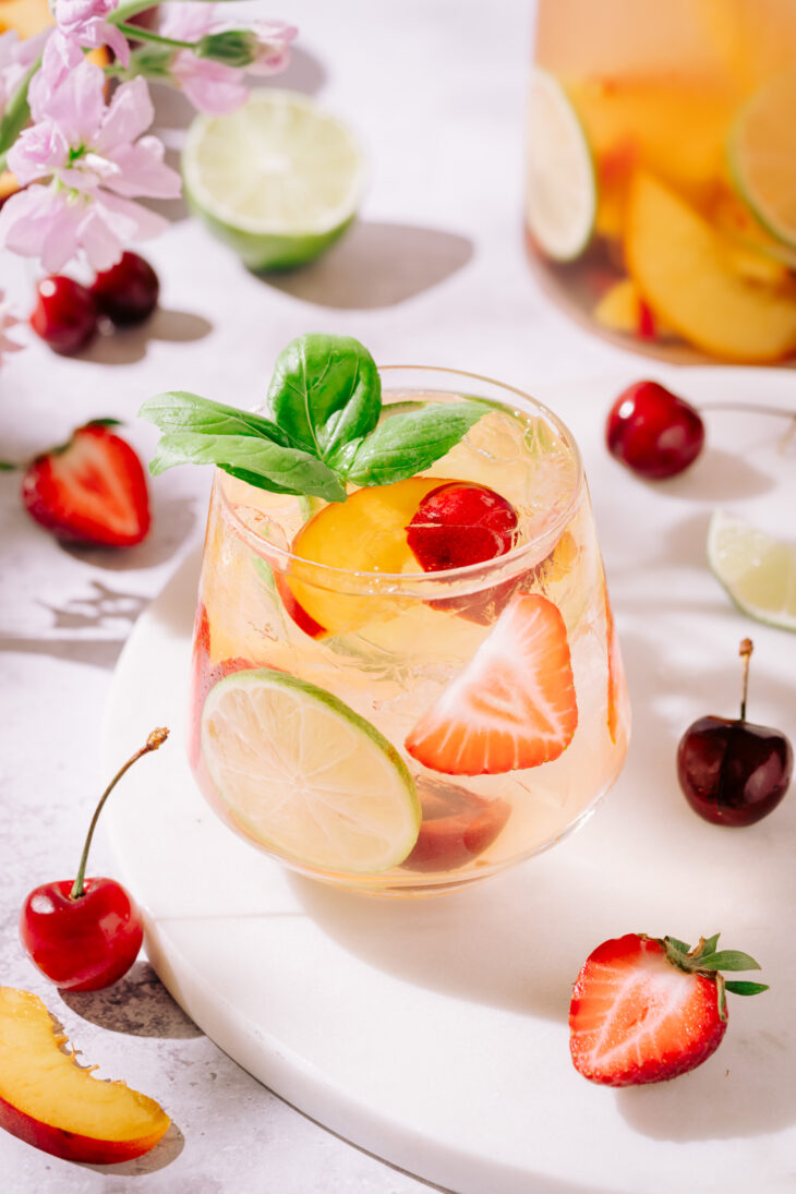 Refreshing summer white sangria in a glass garnished with fresh basil, lime slices, peach wedges, strawberries, and cherries, surrounded by scattered fruits and flowers on a light background.