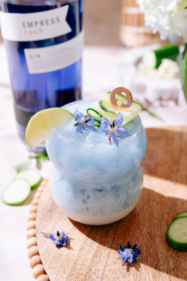 A vibrant blue cocktail named “Cute as a Cucumber” served in a double-bulb glass filled with crushed ice, garnished with a lime wedge, a curled cucumber ribbon, and delicate purple edible flowers, sitting on a wooden board with scattered cucumber slices and flowers.