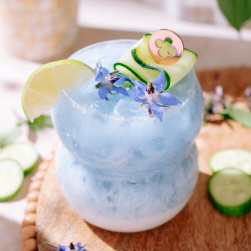 A vibrant blue cocktail named “Cute as a Cucumber” served in a double-bulb glass filled with crushed ice, garnished with a lime wedge, a curled cucumber ribbon, and delicate purple edible flowers, sitting on a wooden board with scattered cucumber slices and flowers.