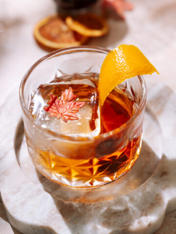 Maple Old Fashioned cocktail in a crystal glass, garnished with an orange peel and maple leaf ice cube, set on a marble coaster.