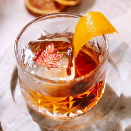 Maple Old Fashioned cocktail in a crystal glass, garnished with an orange peel and maple leaf ice cube, set on a marble coaster.