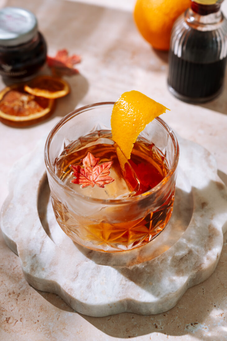 Maple Old Fashioned cocktail in a crystal glass, garnished with an orange peel and maple leaf ice cube, set on a marble coaster.