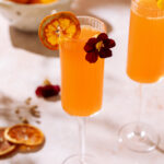 Two Aperol Mimosas in champagne flutes garnished with dried orange slices and edible flowers on a sunlit table
