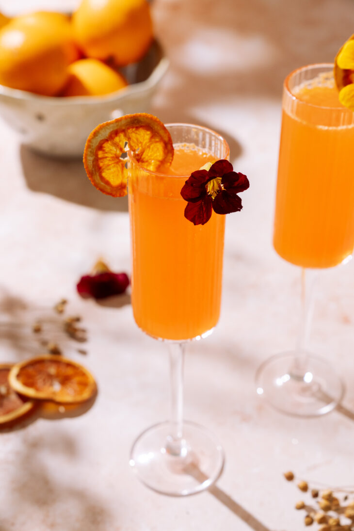 Two Aperol Mimosas in champagne flutes garnished with dried orange slices and edible flowers on a sunlit table