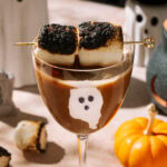 Halloween-themed espresso mocktail garnished with toasted marshmallows and decorated with a ghost design on the glass, surrounded by pumpkins and spooky decor.