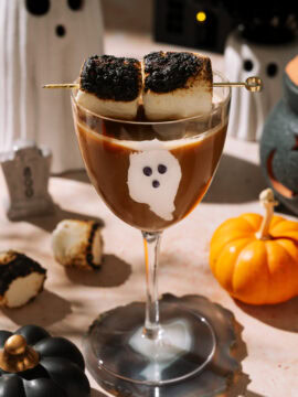 Halloween-themed espresso mocktail garnished with toasted marshmallows and decorated with a ghost design on the glass, surrounded by pumpkins and spooky decor.