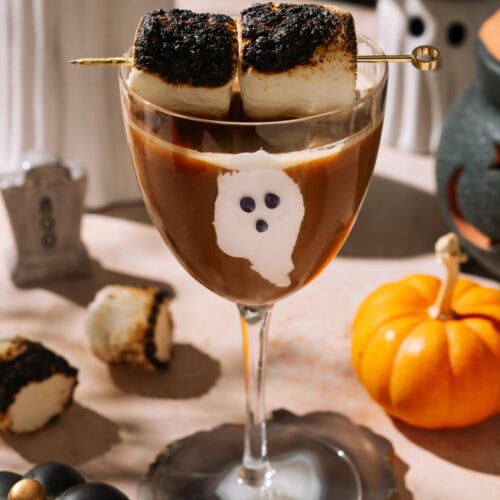 Halloween-themed espresso mocktail garnished with toasted marshmallows and decorated with a ghost design on the glass, surrounded by pumpkins and spooky decor.
