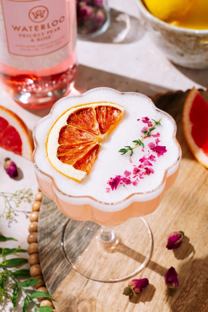 A pink Desert Rose Sour cocktail topped with frothy foam, dried grapefruit slice, and pink flower petals, served in a scalloped coupe glass on a wooden tray. In the background are a bottle of Waterloo Prickly Pear & Rose gin, lemons, and grapefruit slices.