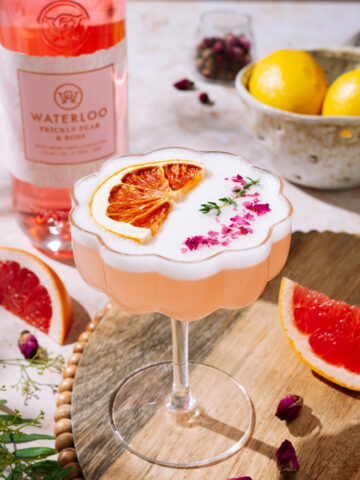 A pink Desert Rose Sour cocktail topped with frothy foam, dried grapefruit slice, and pink flower petals, served in a scalloped coupe glass on a wooden tray. In the background are a bottle of Waterloo Prickly Pear & Rose gin, lemons, and grapefruit slices.