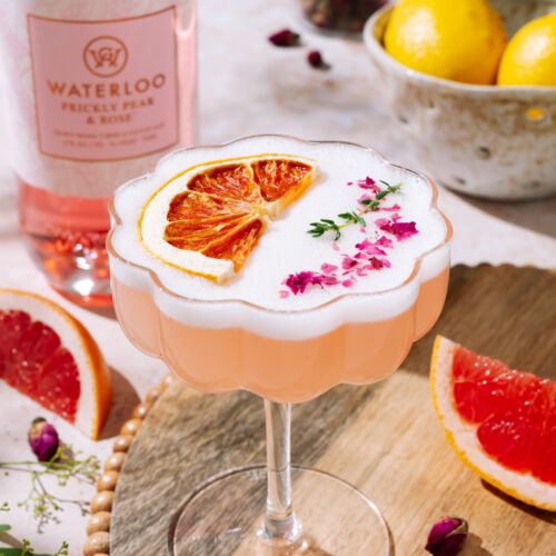 A pink Desert Rose Sour cocktail topped with frothy foam, dried grapefruit slice, and pink flower petals, served in a scalloped coupe glass on a wooden tray. In the background are a bottle of Waterloo Prickly Pear & Rose gin, lemons, and grapefruit slices.