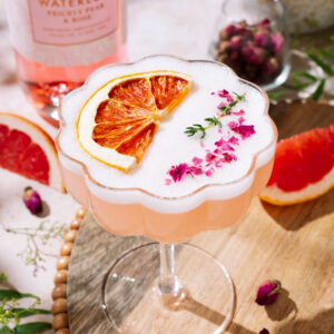 A pink Desert Rose Sour cocktail topped with frothy foam, dried grapefruit slice, and pink flower petals, served in a scalloped coupe glass on a wooden tray. In the background are a bottle of Waterloo Prickly Pear & Rose gin, lemons, and grapefruit slices.