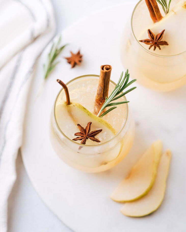 Spiced Pear Vodka Cocktail in a short glass garnished with a slice of a pear, anise, cinnamon stick and rosemary branch. In the background there is one more drink of the same description and 2 pears.