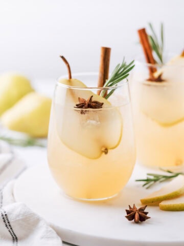 Spiced Pear Vodka Cocktail in a short glass garnished with a slice of a pear, anise, cinnamon stick and rosemary branch. In the background there is one more drink of the same description and 2 pears.