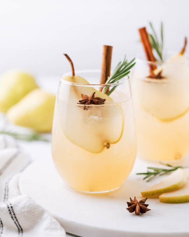 Spiced Pear Vodka Cocktail in a short glass garnished with a slice of a pear, anise, cinnamon stick and rosemary branch. In the background there is one more drink of the same description and 2 pears.