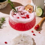Pink Frostbite Christmas Margarita garnished with rosemary, pomegranate seeds, and dried lime.