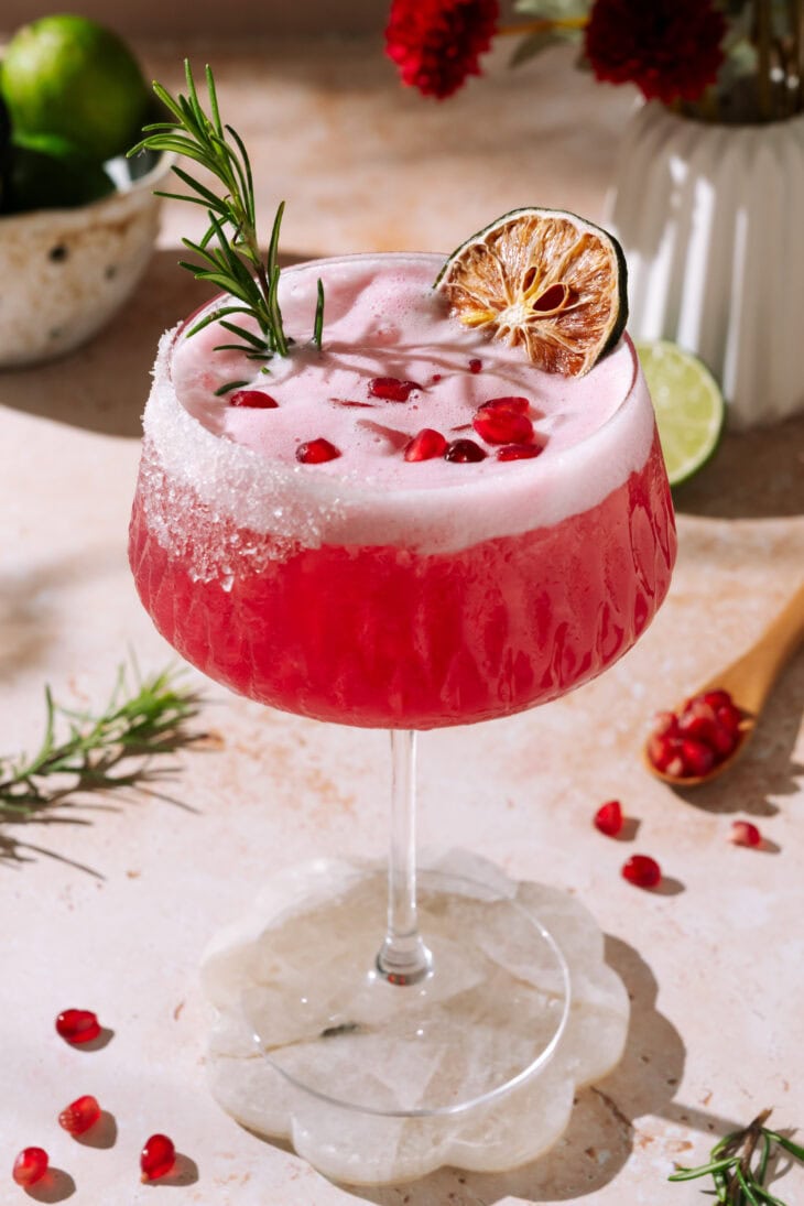 Pink Frostbite Christmas Margarita garnished with rosemary, pomegranate seeds, and dried lime.