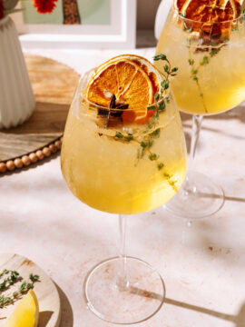 Two refreshing Amaretto Spritz cocktails served in large wine glasses with ice, garnished with dried orange slices, thyme sprigs, and star anise, set on a sunlit table.