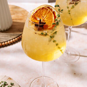Two refreshing Amaretto Spritz cocktails served in large wine glasses with ice, garnished with dried orange slices, thyme sprigs, and star anise, set on a sunlit table.