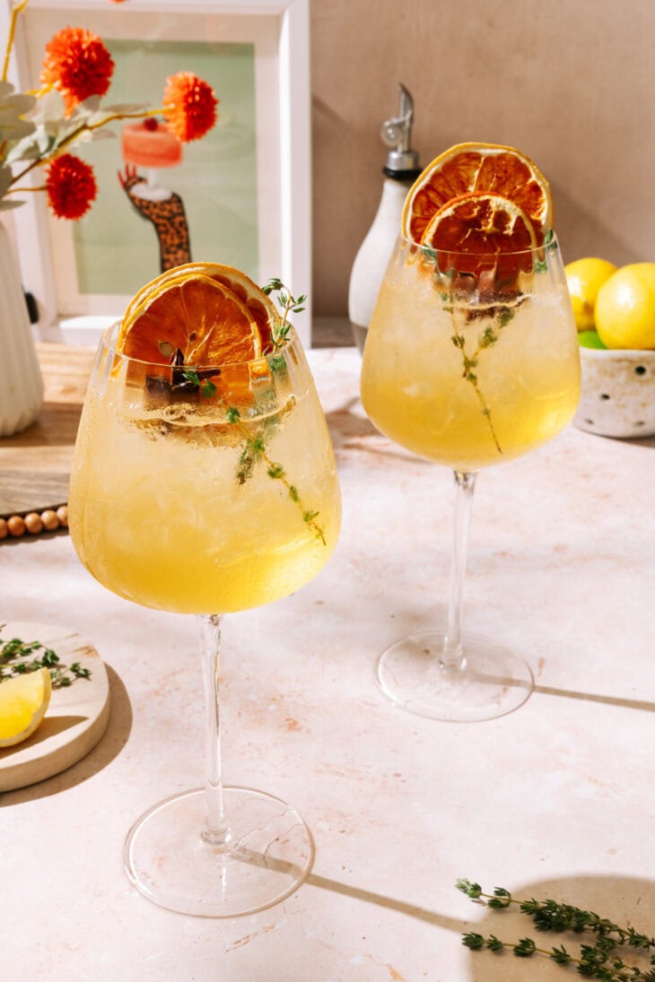 Two refreshing Amaretto Spritz cocktails served in large wine glasses with ice, garnished with dried orange slices, thyme sprigs, and star anise, set on a sunlit table.