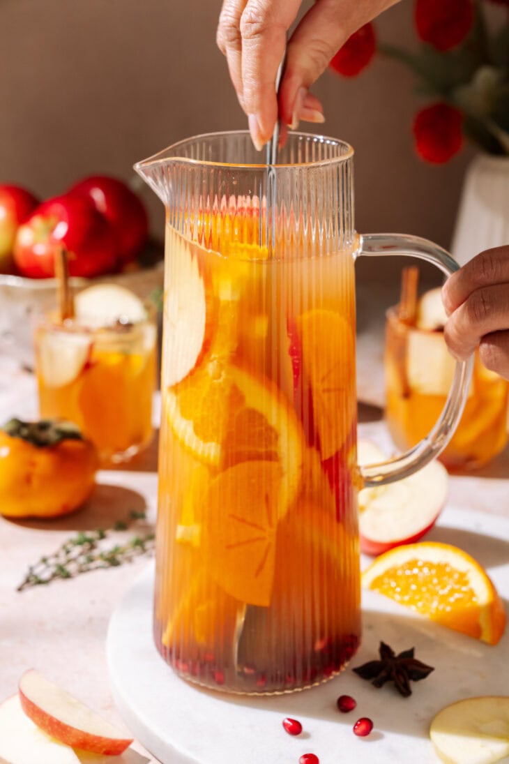 Glass pitcher filled with apple cider sangria and sliced apples and oranges, being stirred.