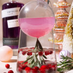 Holiday “Cosmo in the Snow” cocktail served in a clear ornament over a glass with cranberries, rosemary, and sparkling mixer.
