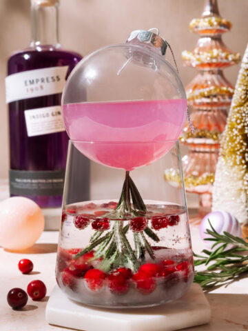 Holiday “Cosmo in the Snow” cocktail served in a clear ornament over a glass with cranberries, rosemary, and sparkling mixer.