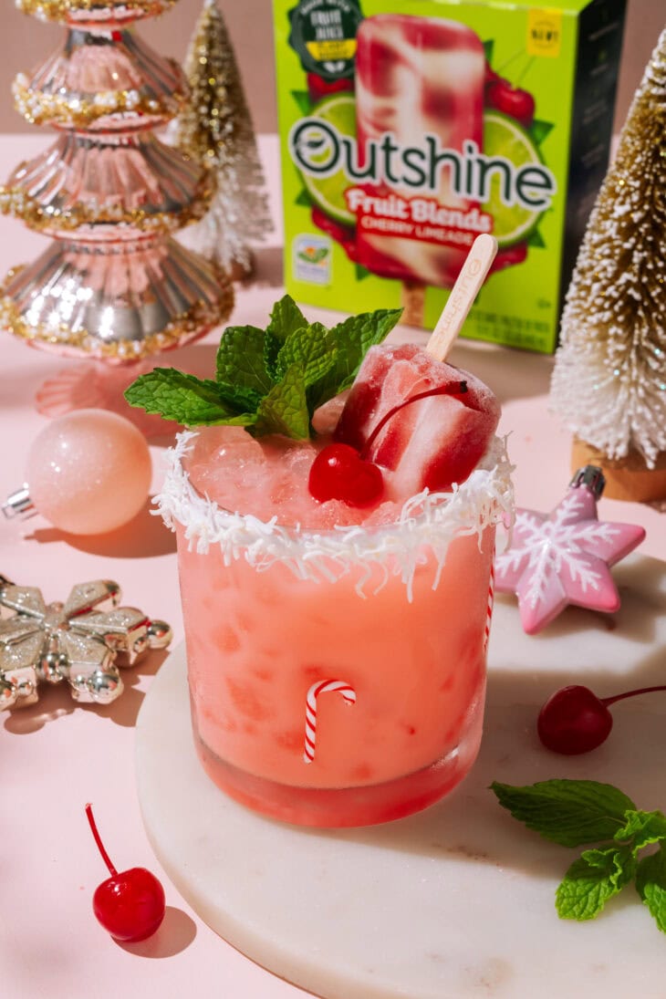Cherry Merry Mocktail with popsicle garnish and mint, styled with Christmas ornaments and an Outshine cherry limeade box in the background.