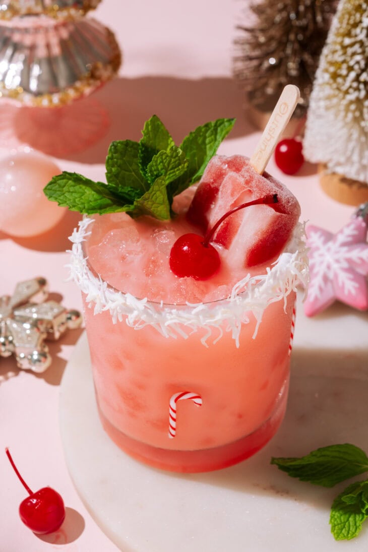 Close-up of the Cherry Merry Mocktail with a popsicle, mint garnish, and coconut-rimmed glass in a pink holiday setting.