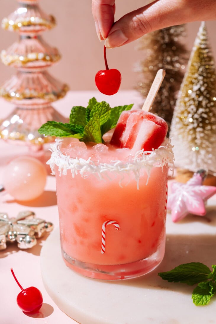 Hand placing a cherry over a festive Cherry Merry Mocktail with a popsicle, mint, and coconut-rimmed glass in a holiday setting.